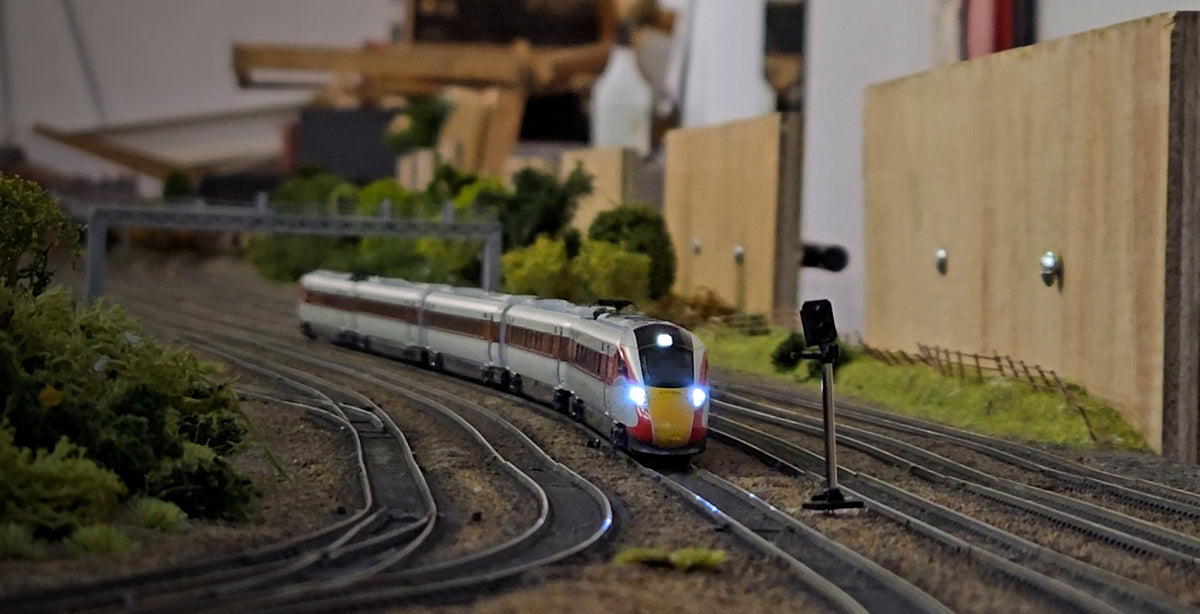 Modern passenger train approaching through a multi-track model railway layout with signals, realistic scenery and detailed trackwork.